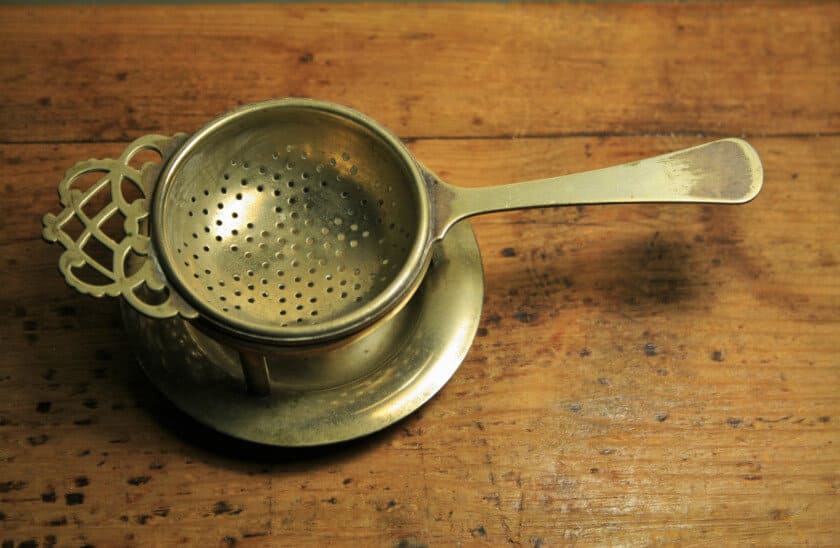 A brass strainer on a stand: strainers are tea accessories used to catch the tea leaves and dust as tea is poured into the cup.