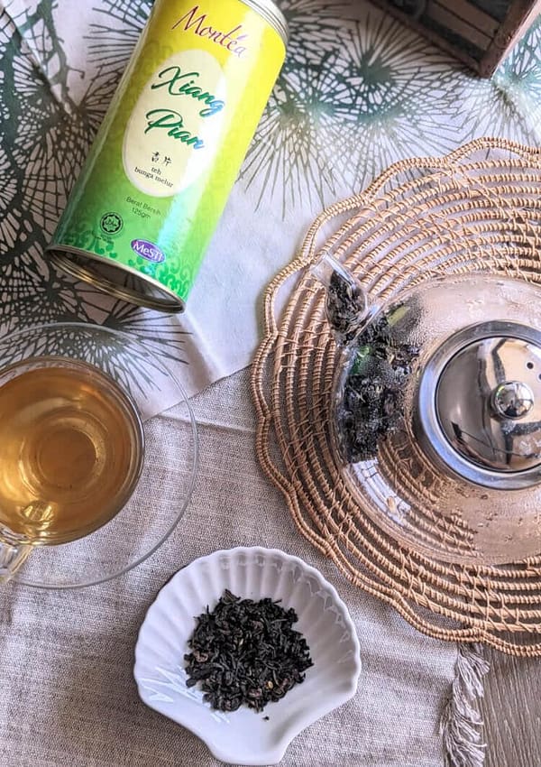 montea xiang pian jasmine tea tin next to a cup of jasmine green tea and jasmine green tea leaves