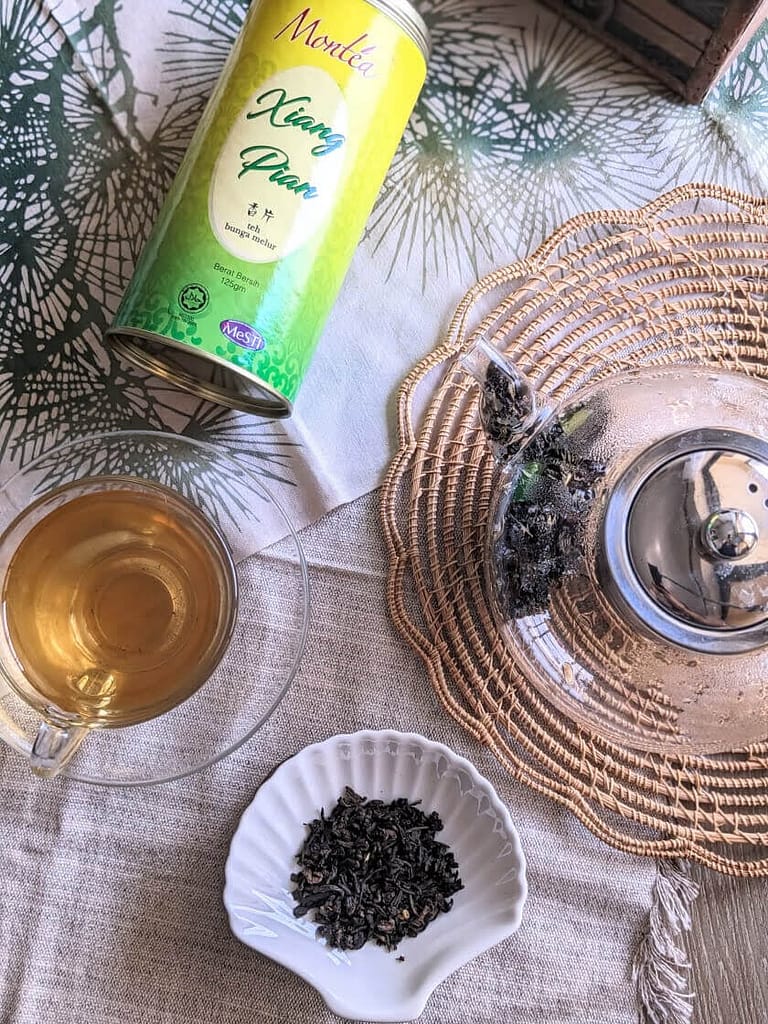 montea xiang pian jasmine tea tin next to a cup of jasmine green tea and jasmine green tea leaves